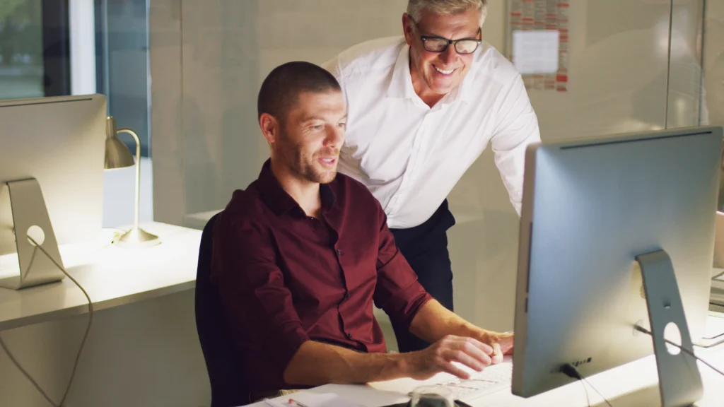 Zwei Männer arbeiten gemeinsam an einem Computer im Büro, einer sitzt am Schreibtisch und bedient die Tastatur, während der andere hinter ihm steht und auf den Bildschirm schaut